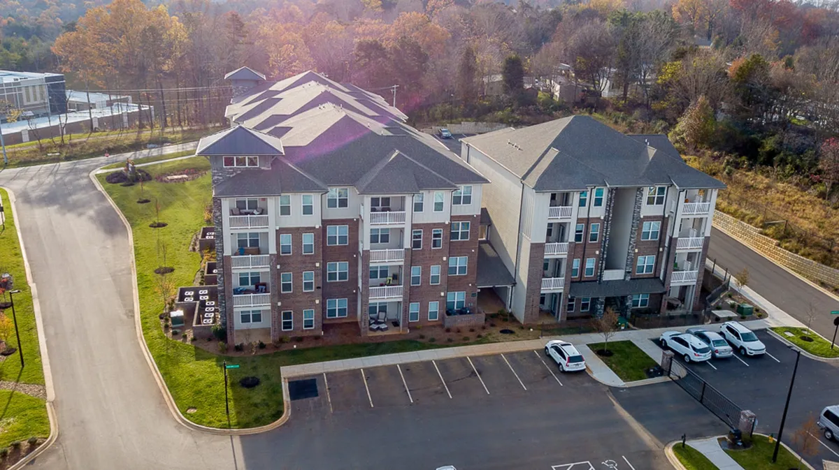 This image presents another angle of the apartment exterior, with a focus on landscaped areas, shaded walking paths, and green spaces for outdoor relaxation or socializing. The well-maintained grounds and nearby trees offer a serene atmosphere, ideal for evening strolls or enjoying nature close to home.