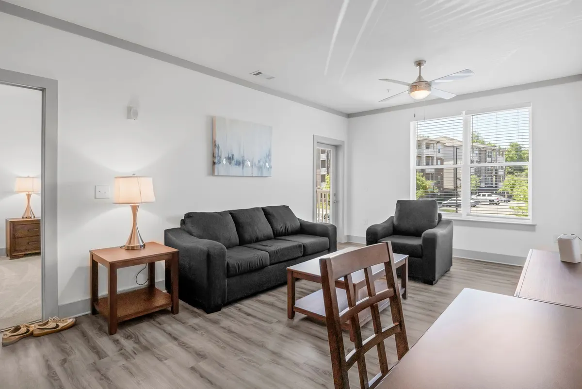 A spacious, chic living area featuring a stylish sofa, modern coffee table, curated artwork, and large windows that fill the space with natural light. The arrangement and decor create an immediate sense of welcome and comfort.