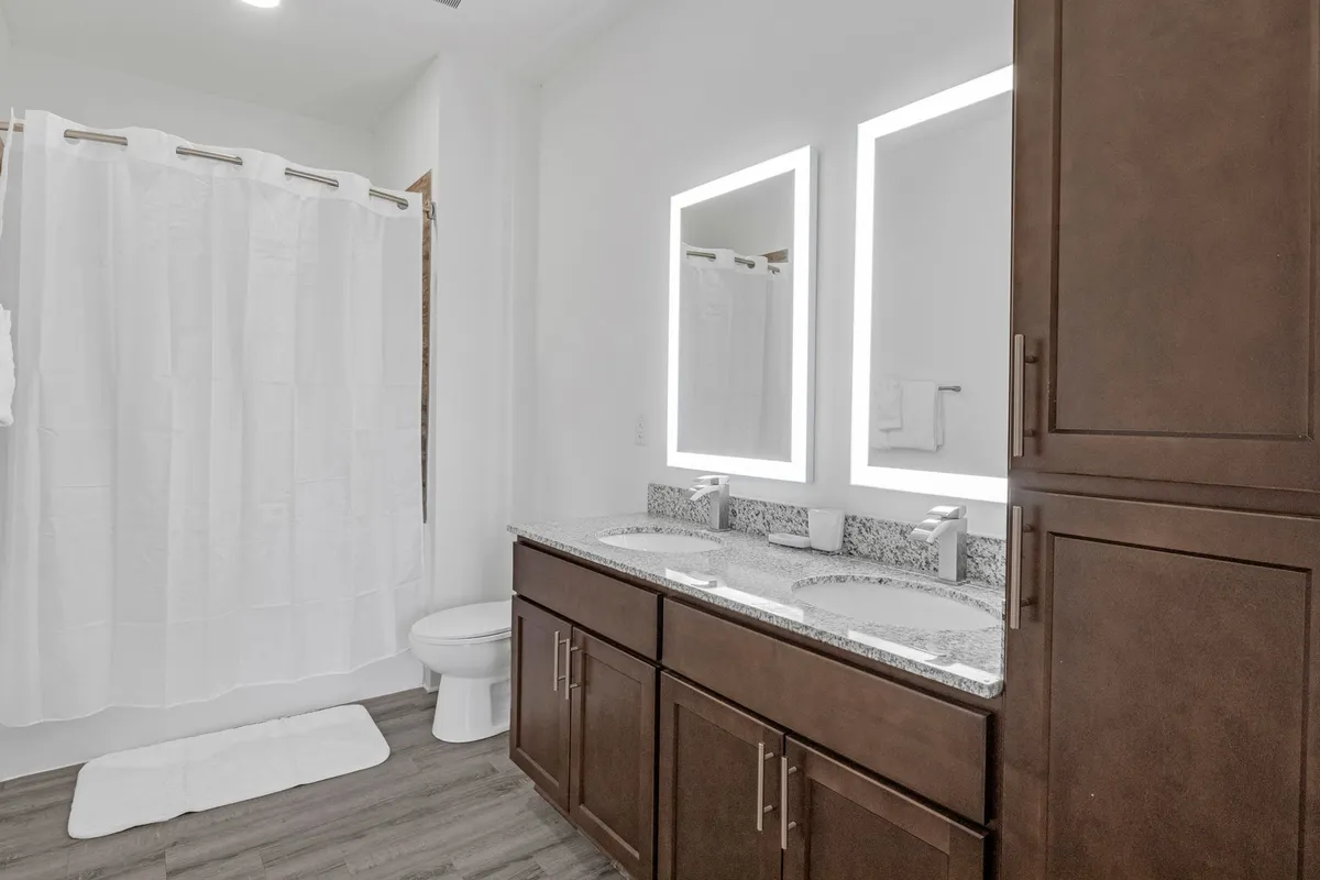 A tranquil bathroom featuring a double vanity, backlit mirrors, granite countertops, and a subtle glimpse of the shower area, all bathed in refreshing natural light. The scene suggests relaxation and spa-like comfort.
