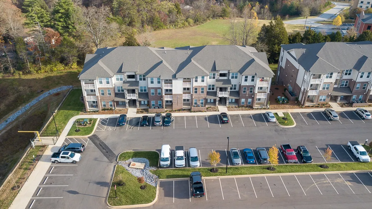 This image captures the exterior of the apartment building, with a landscaped parking lot, mature trees, and inviting green spaces. The image shows residents enjoying outdoor spaces and walking in the neighborhood, conveying ease of access to local amenities and a sense of connection to the surrounding community.