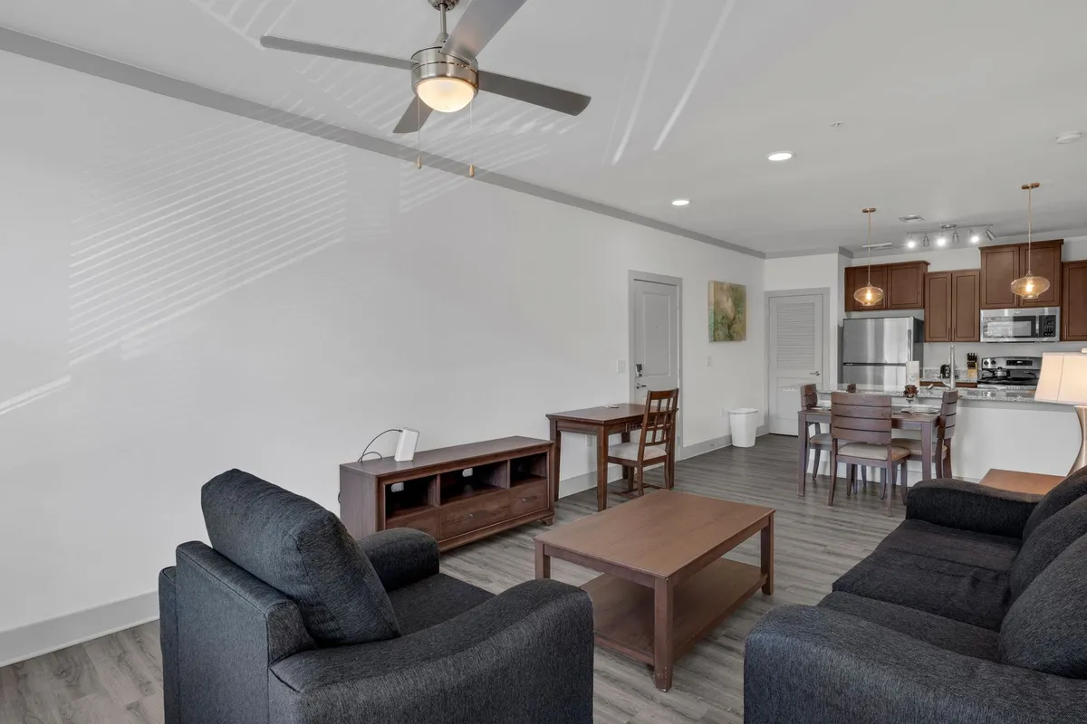 An inviting open-plan living area featuring a plush sofa and coffee table seamlessly integrated with a dining table and kitchen area in the background. Large windows flood the space with natural light, enhancing the contemporary decor and overall comfort.
