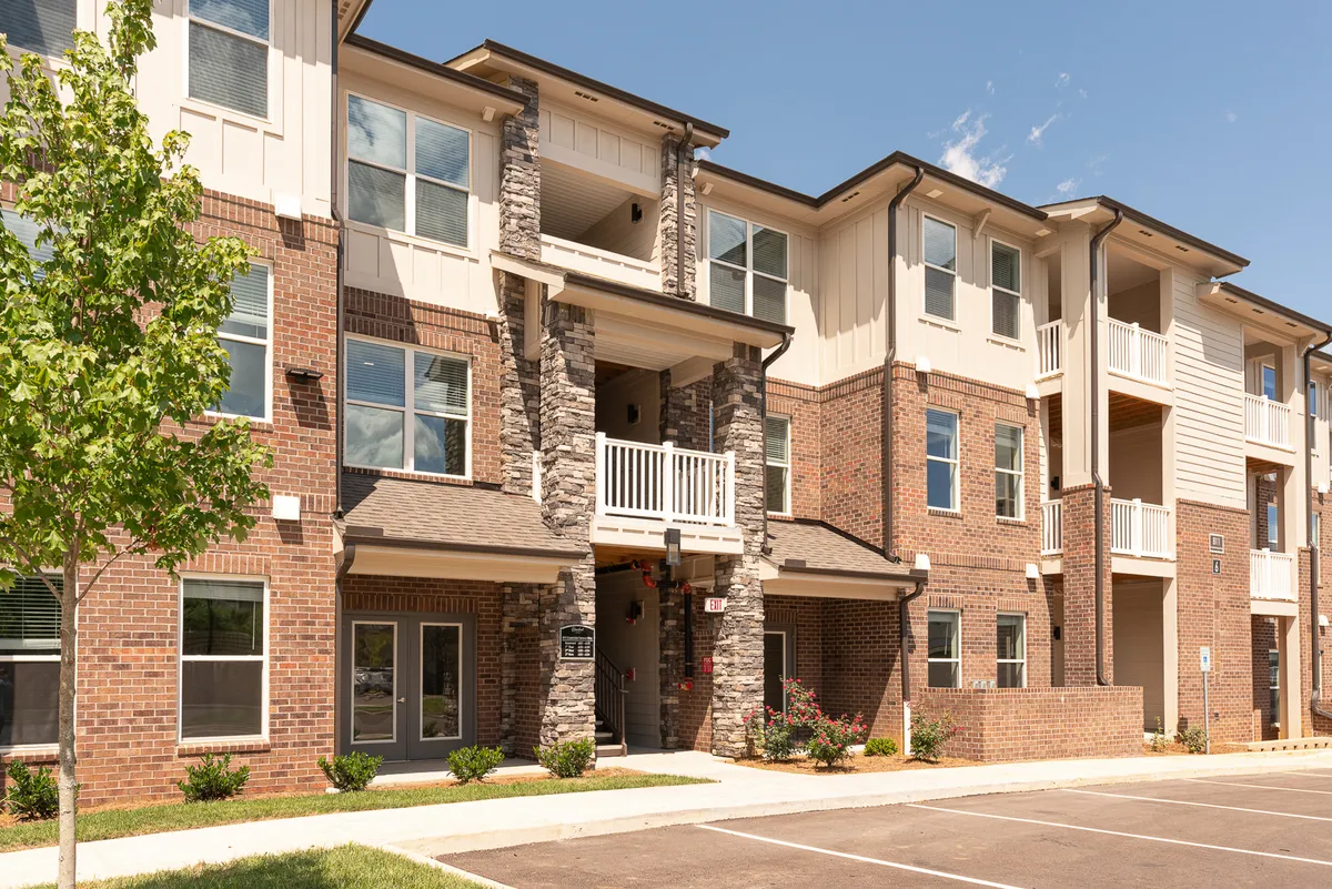 This exterior image showcases the apartment building's stone columns, balconies, large windows, and lush landscaped greenery. The inviting atmosphere is enhanced by the view of balconies perfect for morning coffee or evening relaxation, and the walkable garden pathways hint at the surrounding vibrant community.