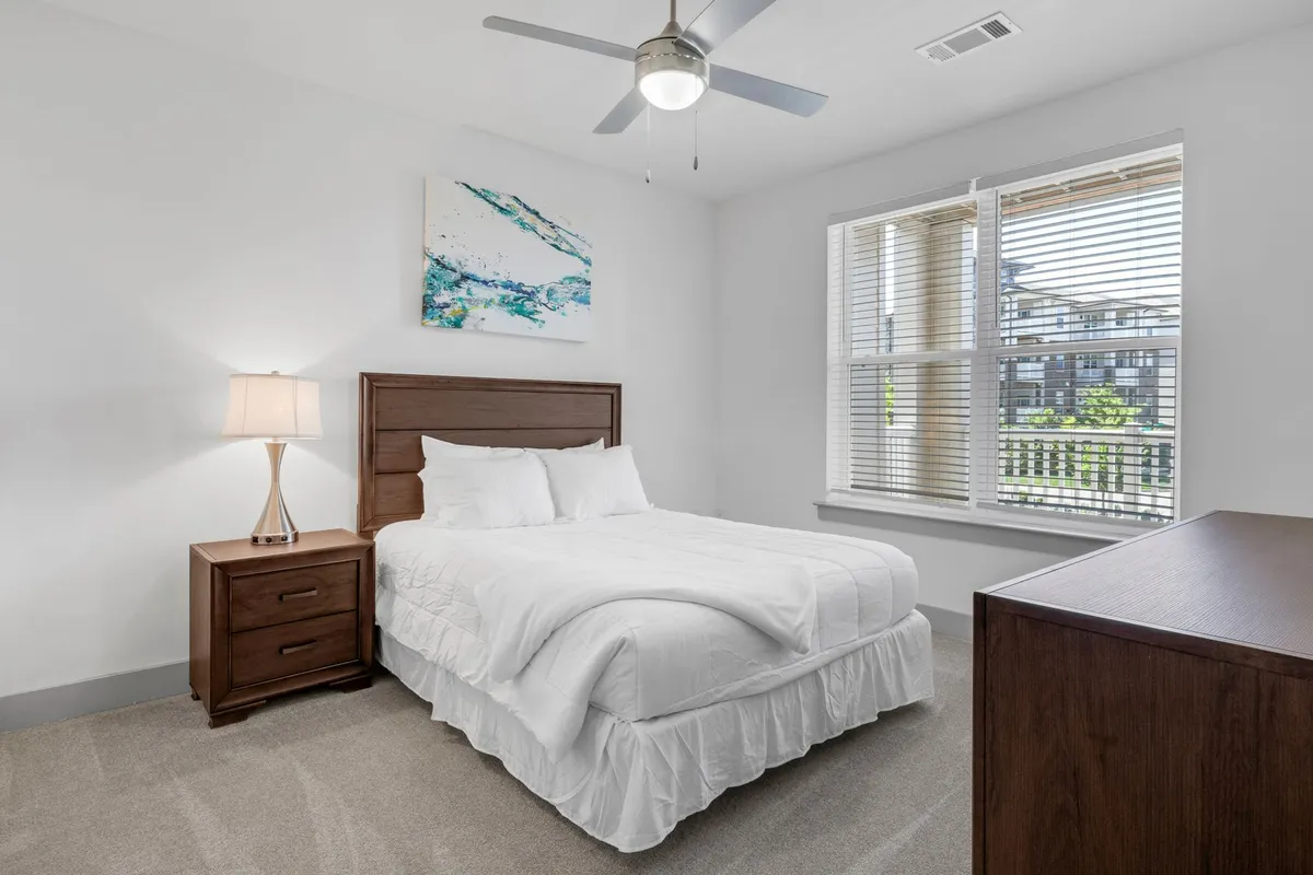 The image showcases a serene, light-filled bedroom featuring a plush bed with crisp linens, stylish nightstand with a reading lamp, modern artwork above the bed, and a large window that bathes the space in natural sunlight. The overall mood is peaceful and inviting, with an emphasis on comfort and relaxation.