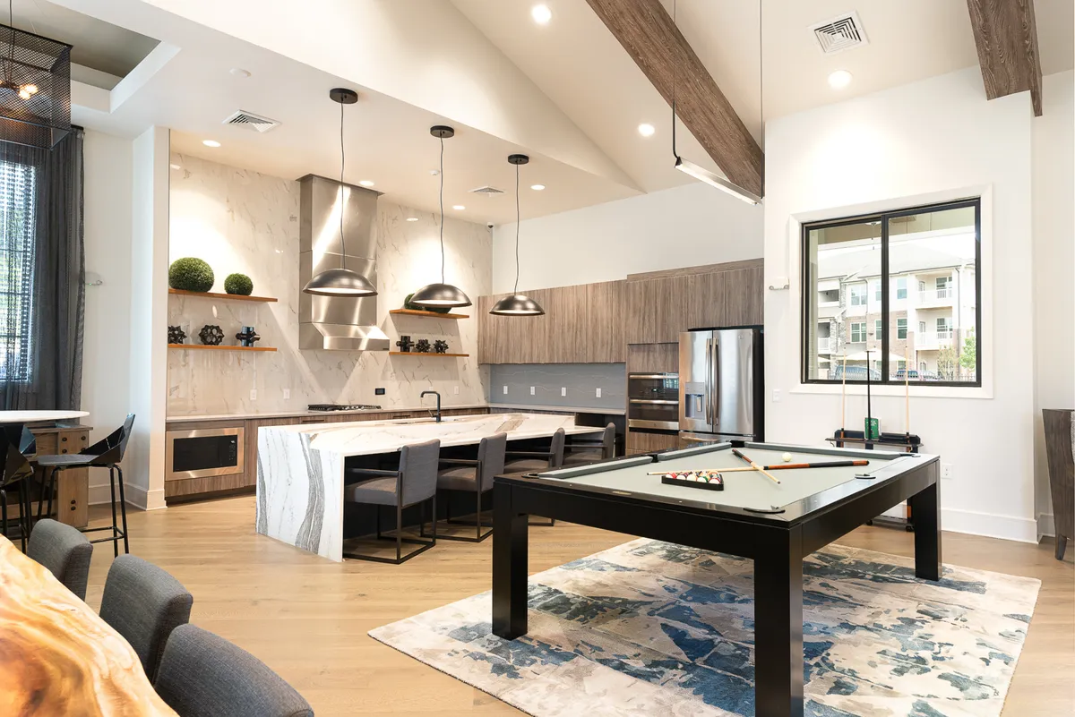 A modern, social space with a pool table, kitchen island with seating, pendant lighting, and large windows that bathe the room in daylight. The arrangement encourages both relaxation and entertaining, echoing a versatile, lively lounge environment.